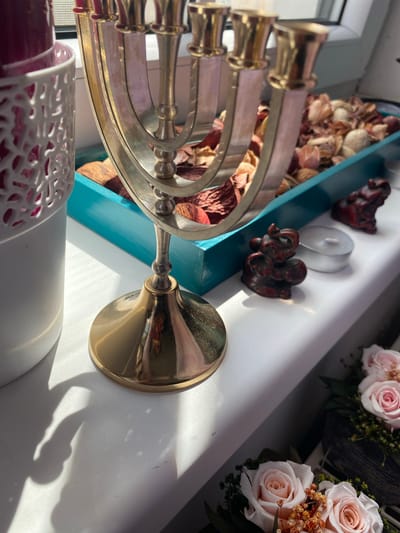 A close-up, eye-level shot focuses on a menorah and decorative items placed on a white windowsill. The menorah, made of a gold-toned metal, stands prominently in the center, with its nine candle holders visible. To the left of the menorah is a white decorative container holding a dark pink candle. Behind the menorah, a teal-colored tray holds dried potpourri. A small, dark red elephant figurine and a tea light candle are positioned to the right of the menorah. In the lower right corner, a bouquet of pale pink roses adds a soft floral element. Sunlight streams in from the right, casting shadows across the windowsill. The overall scene is indoors, likely in a home setting, with a window to the rear. The location is Osek, Czechia.