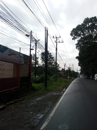 The image depicts a roadside scene in Kamji Na-Yekha, Bhutan, under an overcast sky. A dark, wet asphalt road, marked with a solid white line on its right edge, occupies the right side of the frame, curving slightly into the distance where multiple vehicles, including a white van, are discernible. To the immediate left of the road, a dark, wet gravel and dirt shoulder transitions to a low stone wall. Above this, an orange-brown metal fence with a diamond-grid pattern partially obscures a light-colored building wall. A white, blue, and red banner displaying the text "Decoplus" is draped over this wall and fence. Numerous dark utility poles, heavily laden with extensive networks of overhead electrical and communication wires, extend from the left foreground towards the midground. One pole in the left midground is equipped with a street light. Dense green foliage and trees are visible behind and between the utility poles on the left. Large, mature trees with dark green canopies line the right side of the road in the midground and background. No individuals or specific actions are visible within the frame.