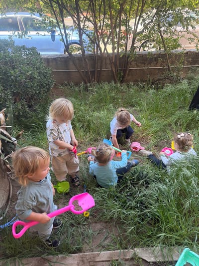 Five young children are playing in a grassy area next to a building. They are using plastic shovels and toys to dig in the dirt. A car is parked nearby, visible through some trees. The specific location is Kiryat Motzkin, Israel.