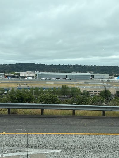 The image shows an aerial view of a Boeing factory in Seattle, Washington, United States of America. The factory is located near a runway and several airplanes are visible on the tarmac. The sky is overcast with clouds. The foreground of the image shows a highway with guardrails and road markings. The overall scene depicts an active aerospace manufacturing and flight testing facility.