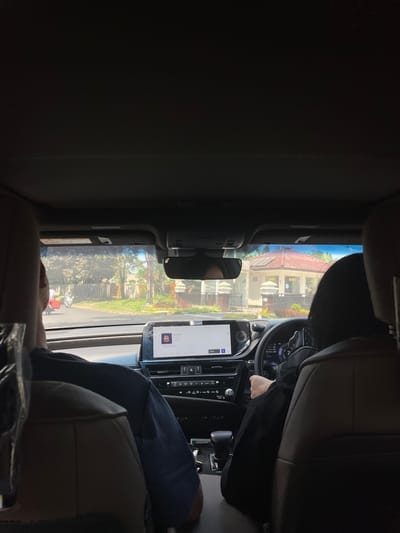 The image depicts an interior view from the rear seat of a vehicle, looking forward through the windshield in the city of Kayuambon, Indonesia, during daylight hours. Two individuals occupy the front seats: a passenger on the left wearing a dark blue shirt, and a driver on the right with dark hair, wearing a black long-sleeved garment, with their right hand positioned on the steering wheel. The car's interior features a dark overhead lining. The dashboard includes a prominent central digital display currently showing a music interface with album art and the title "Hidden Games." Below the screen are various climate controls and buttons. A gear selector is visible in the center console, and the driver's instrument cluster displays "24" (likely degrees Celsius) and "1709.5" (likely kilometers).

Through the windshield, a paved road is visible ahead. On the left side of the road, a person on a red scooter/motorcycle is partially visible, along with another motorcycle further down. Dense tropical trees and green foliage line both sides of the street. On the right side of the road, a light-colored security building with a distinct terracotta-tiled conical roof and "SECURITY" signage is prominently featured, flanked by pillars and a gate. A clear, bright blue sky is visible above the external environment.