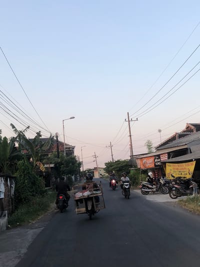 The image depicts a street scene in Glagah Arum, Indonesia, with a gentle twilight sky overhead. Several motorcycles are present on the paved road. In the foreground, a motorcycle is being ridden by an individual wearing a black jacket and helmet, carrying a wooden crate filled with items on the back. To the left, another motorcycle is occupied by a person in dark clothing. Further up the road, other motorcyclists are visible, some riding in groups. The sides of the street are lined with buildings, including structures with tiled roofs and houses with distinct architectural features. Vegetation, such as banana trees and other greenery, is interspersed throughout the scene. Additionally, various signs and banners are displayed, some with text indicating food vendors or businesses. Power lines crisscross above the street, supported by utility poles. The overall impression is of a typical evening in a residential or commercial area of Glagah Arum.