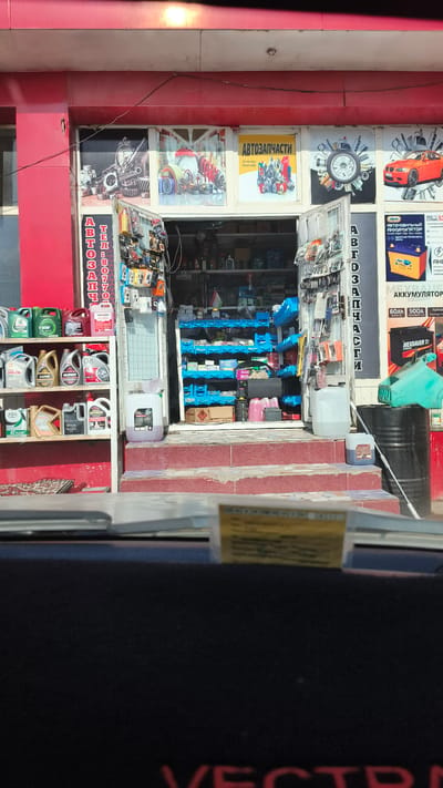 The image shows the exterior of an auto parts store in Mirzo Tursunzoda, Tajikistan. The store is red and white with large windows displaying various auto parts and accessories. The entrance is open, revealing shelves stocked with items. On the right side of the entrance, there is a poster advertising car batteries. The store is situated on a street, with a portion of a car's dashboard visible in the foreground. The scene is well-lit, suggesting it is daytime.