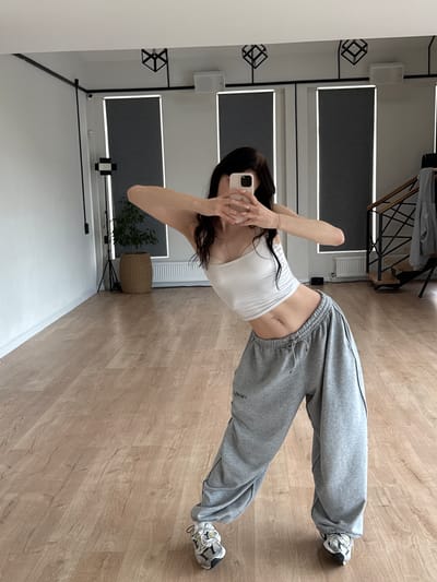A young woman with dark hair is posing in a dance studio, holding a smartphone in her hands to take a mirror selfie. She is wearing a white crop top and loose grey sweatpants, with athletic shoes on her feet. The studio has a wooden floor, white walls, and large windows with grey roller blinds. The background shows a plant in a woven pot, a speaker, and a staircase with a railing. The lighting appears to be bright and natural. The overall scene suggests a practice or rehearsal space.