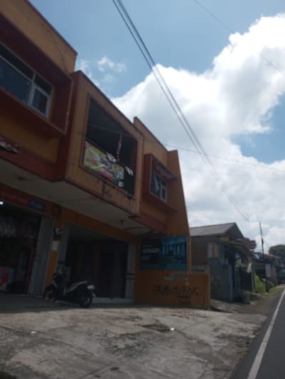 The image shows a blurred street scene in what appears to be a tropical or subtropical region. A two-story building with a brown and tan facade dominates the left side of the frame. The building has windows, signs, and a covered entryway on the ground floor, where a parked motorcycle is visible. To the right of the building, a smaller structure with a corrugated metal roof is present, followed by a street with a white line marking the edge. Utility wires crisscross the sky, which is partly cloudy with a blue sky visible between the clouds. The location is suggested to be Kioumaknao 2, Laos, although this cannot be independently verified from the visual information in the image. The image does not provide information about people or their interactions.