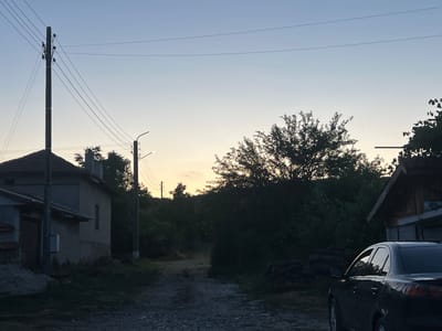 The photograph captures a street scene in the city of Akandzhievo, Bulgaria, during dusk. A dirt road runs through the center, flanked by vegetation and buildings. On the left, a small house with a grey facade and a dark-tiled roof is visible, with a pile of construction debris in front. A utility pole with electrical wires stands beside the house. To the right, a dark-colored sedan is parked. Further down the road, more trees and foliage line the path, and a rustic wooden structure with a sloping roof can be seen in the background. A second utility pole with a street lamp is also present. The sky above is a gradient of pale blue and soft orange, indicative of the setting sun.