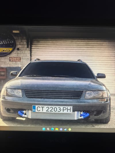 The scene depicts the front exterior of a dark grey Volkswagen Passat B5.5 station wagon parked facing the viewer, positioned in front of a commercial building in Tsenovo, Bulgaria. The vehicle features a custom front-mounted intercooler visible through the lower grille opening, connected by blue silicone hoses. A Bulgarian license plate, "BG CT 2203 PH," is mounted centrally on the front bumper. The car is equipped with roof rails and appears to have tinted windows.
The background consists of a light-colored brick wall building with a large, beige-colored horizontal roll-up metal garage door directly behind the vehicle. To the left of the vehicle, partially obscured by the car, a circular sign is visible on the brick wall, displaying text that includes "Santos" and "TOMS" or "CUSTOMS." Below this, a rectangular sign states "AUTO BODY" and "AUTO PARTS." A red, rectangular object resembling a mailbox or utility box is also affixed to the brick wall on the left. No individuals or discernible actions are present in the image. The environment is illuminated by natural daylight.