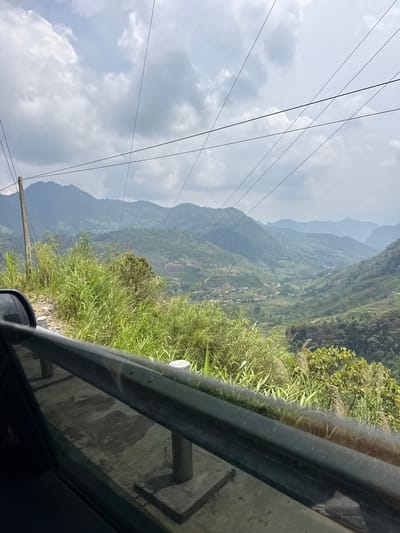 The image presents a scenic view of a vast mountainous landscape, likely captured from a moving vehicle. The scene is composed of successive layers of green-clad mountains and valleys receding into the distance, suggesting an expansive terrain. Lush green vegetation, including tall grass and bushes, dominates the immediate foreground, growing alongside what appears to be a roadside embankment.
A sturdy metal guardrail runs horizontally across the lower right portion of the frame, indicating a road or elevated path from which the photograph was taken. Reflections on a vehicle's window and a partial view of its side mirror are visible in the bottom right corner, confirming the perspective from inside a car or similar transport.
Above the rugged topography, a bright, partially cloudy sky stretches overhead, crisscrossed by multiple dark power lines and supported by a utility pole visible on the left. The clouds are diffused, suggesting a clear daytime, possibly morning or afternoon. The distant mountains appear somewhat hazy, adding depth to the scene. Small clusters of buildings or settlements can be faintly observed scattered within the valleys below, indicating human habitation amidst the natural environment. No people are visible in the image. This serene vista offers a glimpse into a rural, mountainous region, consistent with the provided context of Minh Sơn Commune, Vietnam.