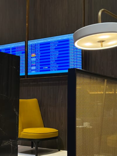 The image shows a departure board at an airport, displaying flight information in Greek and English. The board lists flight times, airlines, flight numbers, destinations (including cities like Skopje, Larnaca, and Tel Aviv), and gate information. A lamp is visible on the left side of the frame. A yellow chair sits next to a table. The departure board indicates a date of 17-05-2026 and a time of 23:40. Based on the destination city names on the board, the location is Spata-Loutsa Municipal Unit, Greece.