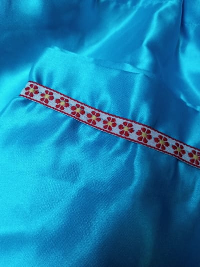 A close-up captures a bright turquoise fabric with a satin sheen. The material is draped, creating soft folds and highlights. A decorative ribbon is attached to the fabric. It has a repeating floral pattern of red petals and yellow centers, set against a white background with a red trim. The focus is on the texture and color contrast.