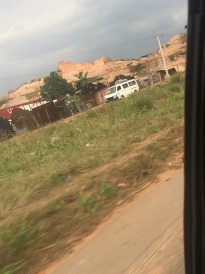 This image captures an outdoor scene in Nwofia, Nigeria, taken from the perspective of a moving vehicle. The foreground consists of a paved road segment in the lower right, transitioning to a reddish-brown dirt shoulder and an unkempt grassy embankment covered in dry and green vegetation. Small scattered debris is visible within the grass.
In the midground, a white commercial van, possibly a minivan or small bus, is stationary, facing left, with items secured to its roof rack. Behind the van and extending to the left, a collection of structures includes several corrugated metal shacks, a prominent red shipping container, and an assortment of metal gates and scrap materials, suggesting a fabrication workshop or junkyard. Sparse green trees and foliage grow amongst these structures.
The background features a large, exposed hillside or cliff face composed of light-brown or reddish-orange earth, indicating an excavated or natural geological formation. Utility poles and power lines traverse the scene. The sky is overcast with heavy grey clouds. No discernible human figures are present. The overall lighting is diffuse, consistent with an overcast daytime environment.
