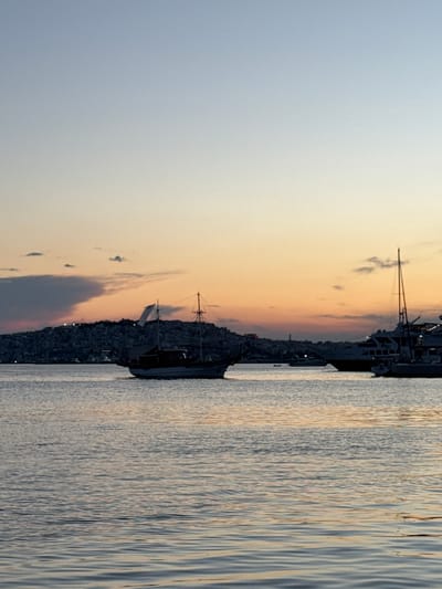 The image captures a serene harbor scene at dusk, with the Municipal Unit of Dafni, Greece, visible as a silhouette of buildings on a hill in the background. The sky transitions from a soft blue at the top to warm hues of orange and yellow near the horizon, suggesting the end of a day.

Several boats are present in the water. In the foreground, the calm water reflects the sky and the surrounding elements, creating a peaceful ambiance. In the mid-ground, a traditional wooden boat with two masts and a white sail is prominently featured. Further back, larger, modern vessels, likely ferries or yachts, are anchored or docked, their lights beginning to twinkle against the fading daylight. The city lights on the hill add a touch of urban life to the natural landscape.

The overall impression is one of tranquility and a picturesque view of a coastal city as night approaches. There are no visible people in the image.