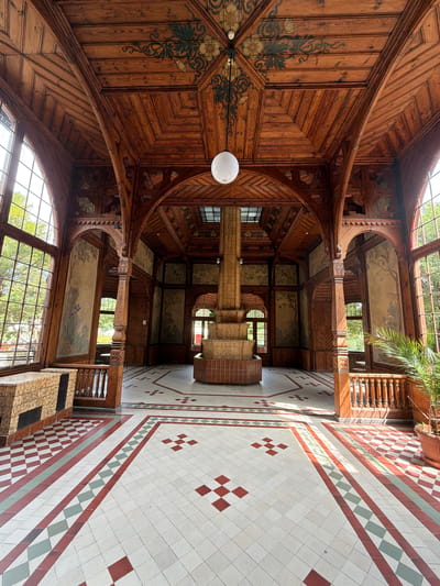 The image shows the interior of the "Czerniańskie Gród" building in Świeradów-Zdrój, Poland. The room features intricate wooden architecture, including a vaulted ceiling with decorative carvings and paintings. A central, multi-tiered brick structure stands in the middle of the room. Large windows with leaded panes line the walls, letting in natural light that casts long shadows on the patterned tiled floor. The floor itself is a mosaic of white, red, and green tiles arranged in geometric patterns. A potted palm plant is visible on the right side of the image. The scene appears to be an empty, public indoor space, likely a hall or atrium, with no people present.