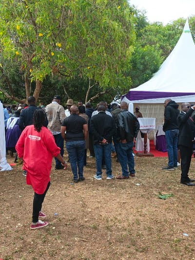 A gathering of people stands on a patch of land with sparse grass, underneath a tree bearing yellow flowers. A white tent with purple trim is visible in the background. The crowd is mostly composed of men, dressed in casual clothing. A woman in a red shirt, labeled "Kone Hall," is walking away from the group. A small podium with a white cloth is visible near the tent.