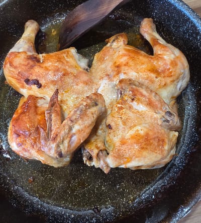 The image shows a roasted chicken cut in half and placed in a black frying pan with some cooking oil. A wooden spoon lies near the chicken. The scene appears to be indoors, on a wooden surface. The location in Novachene, Bulgaria is not discernible from the image.