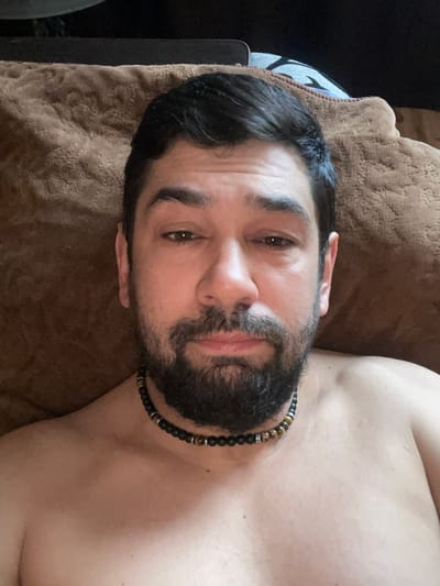 The image is a close-up, selfie-style photograph of a man lying down. He has dark hair, a full beard, and is wearing a beaded necklace. He is shirtless, and the background appears to be a textured brown surface, possibly a blanket or cushion. The lighting is soft and appears to be natural. There is no information in the image to suggest a specific location within the city of Neryungri, Russia.