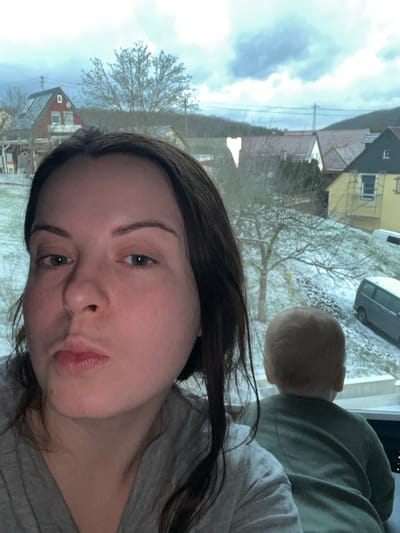 A selfie-style shot features a young woman with dark hair wearing a gray shirt. Her face is the primary focus, capturing her light skin and neutral expression. Outside the window behind her, a snowy landscape includes houses, bare trees, and a vehicle. A baby's head is visible in the upper right corner, along with a green shirt. The photo appears to be taken indoors, possibly near a window overlooking a winter scene.
