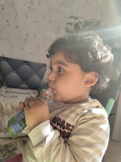 The scene is located in a domestic interior in Карамыш, Russia. A young child, appearing to be a toddler, is depicted in profile view, facing right. The child has dark, curly hair and a light complexion, actively consuming liquid from a green and white flexible pouch with a white spout, holding it with both hands to their mouth. The pouch features cartoon characters and a barcode. The child is dressed in a long-sleeved shirt with alternating beige and white horizontal stripes, displaying partially visible dark lettering, likely "MICKEY", on the chest. The background consists of a light-colored wallpaper patterned with a brown floral or vine motif. To the left, a dark, tufted cushion or upholstered furniture element is visible. A houseplant is present in the upper right background, and a vertical light green object is partially visible on the far right. The environment is illuminated by strong natural light from the right, creating lens flare and bright highlights on the child's hair and the upper right portion of the image.