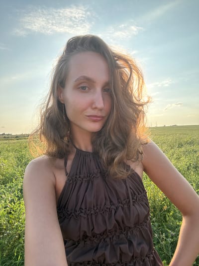 A young woman is taking a selfie in a sunlit field. She has long, wavy brown hair and is wearing a brown, ruffled halter dress. Her left arm is bent, with her hand resting on her hip. The background shows a vast, green field under a bright sky with scattered clouds. The scene appears to be in Aksaitovo, Russia, given the landscape.