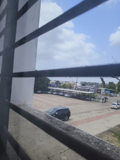 A wide shot, taken from behind a window grate, captures a view of a street and parking area in Dhandhuka, India. The foreground is dominated by a concrete ledge and the dark, angular shadows of the window grate. Beyond this, a paved parking lot with a red and grey patterned surface is visible. Several vehicles are parked in the lot, including a black SUV, a white hatchback, and numerous motorcycles under a covered carport. In the background, a street runs parallel to the parking area, with a few more vehicles, including a white car and a motorcycle with a rider, on it. Buildings line the street, some with signage indicating commercial establishments. A large tree stands behind the parking lot, and the sky above is partly cloudy.