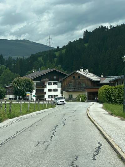 The image depicts a rural road in Reitdorf, Austria, curving through a village with traditional alpine architecture. The road surface is asphalt, showing signs of wear and tear with numerous cracks and dark patches. On the left side of the road, a wooden fence with vertical posts separates the road from a grassy field. Several multi-story buildings, characteristic of Austrian villages, are visible. One prominent building on the left is a white structure with multiple balconies and brown shutters. To its right, a larger building features a dark wood facade with balconies and a steeply pitched roof. In front of these buildings, a white van is parked on the side of the road. Lush green trees and dense evergreen forests cover the rolling hills in the background, leading up to a cloudy sky. A power transmission tower with lines is visible on a distant hill. The overall atmosphere is overcast, suggesting a cool or damp day.