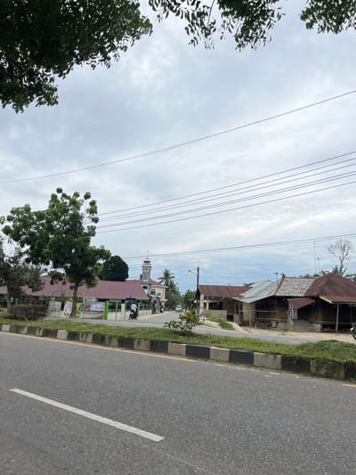 A medium shot captures a street scene in Tanahterban, Indonesia, with a cloudy sky overhead. In the foreground, a paved road with a white dashed line runs diagonally from the bottom left. A concrete curb with alternating black and white sections and a strip of grass separates the road from the sidewalk area. Along the sidewalk, there are several buildings. On the left, a low-rise building with a red roof is visible, possibly a school or community center, with posters displayed on its green fence. A tall, leafy tree stands in front of this building. Further down the street, a minaret topped with a dome and crescent moon rises above other buildings, indicating a mosque. To the right of the mosque, a row of simpler wooden structures with corrugated metal roofs are present. Several power lines crisscross the sky. A motorcycle with two riders is seen on the road in front of the buildings, and a car is visible in the distance on the left. The overall atmosphere appears to be a typical day in a semi-urban or rural Indonesian town.