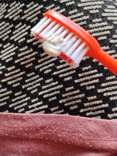 An overhead, close-up shot shows a bright orange toothbrush with white bristles, resting on a black and beige patterned rug. A small amount of white toothpaste is applied to the bristles. In the foreground, a pink fabric with small white flecks lies across the bottom of the frame. The image appears to be taken in a residential setting in Cîrpești, Moldova.