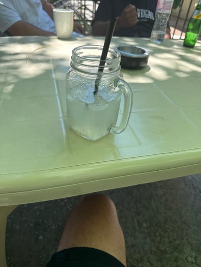 The image depicts a close-up view of a table at an outdoor seating area, likely a cafe or restaurant, in Orizovo, Bulgaria. The table is a light green, tiled surface, with visible grout lines creating a grid pattern. Sunlight casts dappled shadows from overhead foliage onto the table.
In the foreground, a glass mason jar filled with a light-colored beverage and ice cubes is prominently placed. A black straw extends from the drink. To the right of the jar is a dark-colored ashtray.
In the background, out of focus, are parts of individuals and beverages. A white shirt sleeve and shoulder are visible on the left, suggesting someone is seated at the table. Further back, a white mug or glass is present. To the right, a clear plastic bottle of water and a green glass bottle, possibly of soda, are on the table. A portion of a dark t-shirt with the word "VEGAS" visible is also in the background.
The overall impression is that of a relaxed outdoor dining or drinking setting.