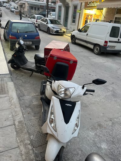 The image shows a white scooter with a red delivery box, parked on the street. The scooter is facing the camera and is situated on the left side of the image. The scooter has a black seat, black handlebars, a black rearview mirror, and a black headlight. The red delivery box is attached to the back of the scooter and has a white label on it. The street is gray and has a sidewalk on the left side. There are several cars parked on the right side of the image, including a white van with the license plate KBP 29974. The scene is set in Limenaria, Greece, at coordinates 40.627, 24.575. A person is walking on the sidewalk to the left. The sidewalk is made of gray concrete. There are two other motorcycles behind the scooter. The street has white markings. A small blue car is parked behind the white scooter. The cars are all parked on the right side of the image. There is a storefront on the right side of the image with a sign that reads "Status". There is a sign in Greek and a sign in English that reads "Cat" above the storefront. There is a building in the background. There are trees and a small white car in the distance. The image was taken during the daytime, but the lighting is somewhat dim. The sky is overcast.