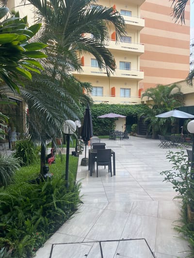 This is an outdoor scene at what appears to be a hotel courtyard. In the foreground, lush green foliage, including palm fronds and ferns, dominates the frame. Behind the greenery, a patio area is visible with light-colored tiled flooring. Several dark-colored outdoor chairs and tables are arranged on the patio, some of which are covered by black and pink umbrellas. In the mid-ground, a building with a beige facade and orange accents is visible, featuring multiple windows. The building's upper section has a distinctive pattern of alternating beige and light orange stripes. To the left of the building, more palm trees and greenery are present. In the background, a taller building with a white facade is partially visible. The overall impression is that of a tropical or semi-tropical environment, likely a resort or hotel, during daylight hours. The lighting suggests it could be morning or late afternoon, and the presence of umbrellas and the lush greenery might indicate a warm and possibly humid climate, consistent with Abidjan, Ivory Coast. No people are visible in the frame. There is no visible text in the image.