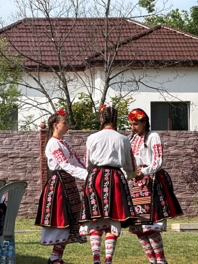 In Pordim, Bulgaria, three young women stand in a semi-circle, facing each other, during an outdoor cultural event. They are dressed in traditional Bulgarian folk costumes. Each costume features a white embroidered blouse with red and black trim, and a layered skirt. The skirts are primarily red and black with intricate floral embroidery and geometric patterns. The women have their hair braided and adorned with flowers. They are positioned in front of a stone wall, with a building with a red-tiled roof and bare trees visible in the background. The ground is covered in green grass, and a plastic chair and bottles of water are present on the left side of the frame. The scene suggests a moment of performance or preparation for a traditional Bulgarian celebration.