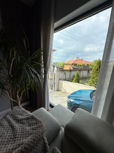The image shows an interior view looking outward through a window. Partially visible outside is a blue car, a section of a building with an orange tile roof, and greenery. The interior contains a gray upholstered couch and a houseplant with spiky leaves. The location is likely in Dolno Drianovo, Bulgaria, but this cannot be definitively confirmed from the image. No people are visible.