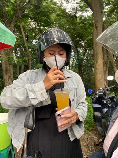 A person wearing a helmet and a white face mask is seen sitting on a scooter or motorcycle in an outdoor setting with lush greenery and trees in the background. The person is wearing a light denim jacket over a black dress. They are holding a clear plastic cup filled with a yellowish liquid, using a black straw to drink from it. A pink-cased cell phone is held in the same hand.