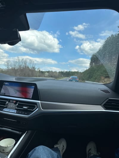 The view from inside a car on a road with a blue sky and scattered clouds. The dashboard of the car is visible, showing a navigation screen with a map and car controls. The driver's legs and white Adidas sneakers are partially visible. A coffee cup with "Bianchi" on it is present in the car. The location is unclear from the provided image; therefore, stating the location to be in Bregovo, Bulgaria, is not possible.