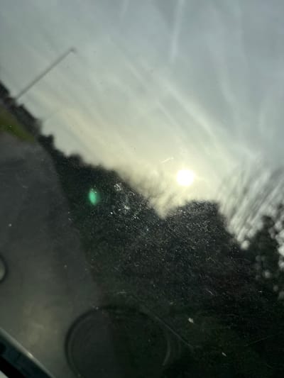 This image captures a static outdoor scene in Baschurch, United Kingdom, viewed through a heavily smudged and dusty glass surface, likely a vehicle windshield. The sky, partially obscured by dirt and glare, appears overcast with bright sunlight originating from the upper right quadrant, producing a distinct green lens flare in the mid-left. A blurred, dark paved surface, possibly a road or path, traverses diagonally from the lower-left to the upper-left. Along the horizon, a dense, dark line of trees or shrubbery is silhouetted against the brighter sky, further obscured by the light source. A slender, dark utility pole is visible in the upper-left, extending upwards against the pale sky. The immediate foreground shows dark, indistinct curved and circular shapes, consistent with interior vehicle components such as a dashboard or speaker grilles. No individuals or active subjects are present.