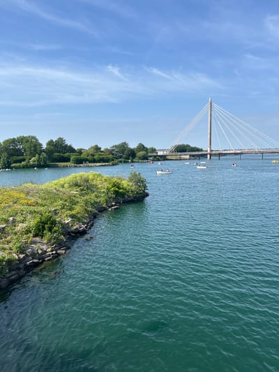 The image depicts a cable-stayed bridge spanning a body of water, likely in Southport, United Kingdom. The single-tower bridge features numerous cables radiating from its apex. The water, possibly a river or a channel, is calm with a slight ripple effect. Several small boats are scattered across the water's surface. A narrow strip of land is visible on the near side of the water, covered with lush green vegetation and rocky embankments. The sky above is clear and blue with scattered wisps of clouds. The perspective of the image is taken from an elevated position, possibly from another bridge or structure.