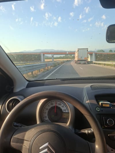 The image is a first-person perspective from inside a car, showing the dashboard and the road ahead. The car appears to be driving on a highway. The steering wheel is visible in the foreground, and a speedometer shows a speed of approximately 100 km/h. A large truck is driving in front of the car. There is an overpass ahead, and a clear blue sky with some clouds. The environment appears to be rural or suburban. The image is taken during daylight, likely in the afternoon, given the warm light. The location is identified as Skravena, Bulgaria.