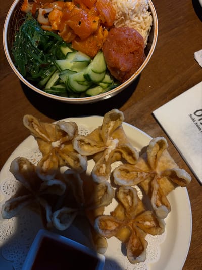 A wooden table presents two plates. One bowl contains rice, salmon, tuna, seaweed salad, and sliced cucumbers. Next to it, a plate holds multiple crab rangoon shaped like flowers, fried until golden, with a small square dish of dipping sauce. A white napkin with dark text sits behind the plate of rangoon.