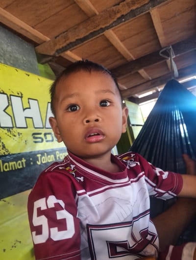 A close-up, medium shot captures a young boy from the chest up, looking directly at the camera. He has dark skin and short black hair. He is wearing a maroon and white sports jersey with the number "53" prominently displayed on the sleeve. The jersey also features a cartoon skull and crossbones emblem on the shoulder. The boy's expression is neutral.

Behind the boy, a yellow banner with large white text is visible. Partially obscured text reads "KHAN", "50", and "lamat : Jalan Por". The background also shows a thatched wooden ceiling with exposed beams and a dark, ribbed structure that appears to be a hammock or draped fabric. Sunlight casts a bright glare on the right side of the image, suggesting it is daytime. The overall setting appears to be outdoors or in a partially covered outdoor area, possibly a home or market stall in Pompanua, Indonesia.