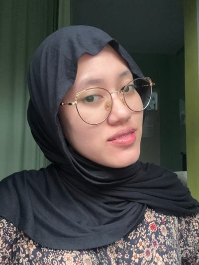 A medium close-up portrait depicts an adult female positioned indoors in Patrang, Indonesia. The individual faces slightly right of the camera, looking directly forward. She wears a dark, solid-colored hijab and gold-framed, circular prescription glasses with decorative scrollwork on the upper outer corners. Her lips display a light pink, glossy cosmetic, and dental braces are visible on her upper teeth. The subject's shoulders reveal a garment patterned with small floral designs in red, white, purple, and green against a dark background. The background is out of focus, featuring a muted green wall or surface, with a partial, indistinct white frame of what appears to be a window or doorway visible on the right side. Illumination appears soft and consistent.