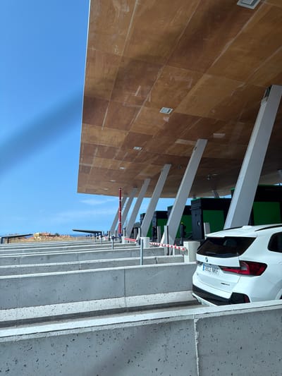 A white SUV, bearing the license plate "0802 NKF," is positioned at a toll booth on a clear, sunny day. The vehicle is facing away from the camera, with its rear clearly visible. The toll booth area is characterized by a modern, open-air design with a large, wood-paneled roof supported by angled white pillars. The roof is tiled with square panels of wood grain material and features integrated square lighting fixtures. In the background, several identical toll booths with green panels and black enclosures are visible, suggesting a toll plaza. Concrete barriers define the lanes, and a red and white striped barrier gate is lowered, indicating that the lane is closed or awaiting a vehicle. The environment is bright and airy, with a vast expanse of blue sky overhead. In the distance, a cluster of buildings with terracotta roofs, characteristic of a Mediterranean coastal town, can be seen against a backdrop of the horizon. This scene is located at the AP-7 toll road in Mijas, Spain.