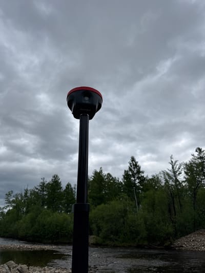 The media file shows a close-up of a black selfie stick with a red suction cup attachment on the end. The selfie stick is angled upwards, with the sky filling most of the frame. The sky is overcast with gray clouds, suggesting a cloudy or rainy day.
In the background, a tree line with lush green foliage is visible along the right side of the image. Beyond the trees, a body of water, likely a river, can be seen with a rocky shore. The water appears dark and reflective, with some ripples on its surface. The overall scene suggests an outdoor setting, possibly in a natural environment like a park or by a river.
The focus is on the selfie stick and its attachment, which is a common accessory for smartphones or cameras used to take photos or videos. There are no people visible in the frame, nor any specific activity taking place beyond the implied use of the selfie stick. The lighting is diffused due to the cloudy sky, and there are no strong shadows. The location is identified as Murtygit, Russia. No visible text is present in the image.