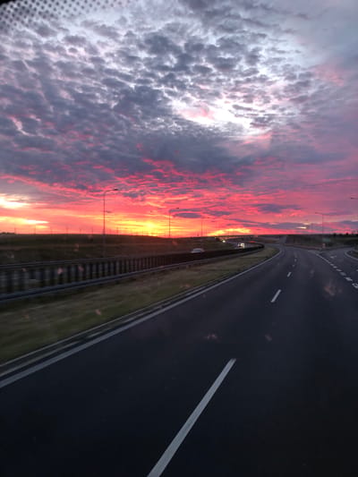 The image shows a dynamic sunset over a highway in Zaparcin, Poland. The sky is ablaze with vibrant shades of pink, orange, and yellow, illuminated by scattered clouds. The highway stretches ahead, with visible lane markings and guardrails. Several vehicles are present on the road, indicating moderate traffic. Streetlights line the sides of the highway, and a grassy embankment is visible to the left of the road. The overall impression is one of a journey nearing its end as day transitions into night, with a spectacular natural display overhead.