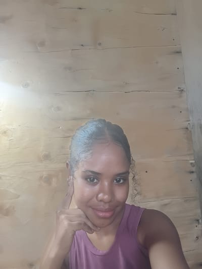 A person with dark hair and skin is facing the camera with a slight smile. They are wearing a mauve top. Their left hand is touching their face. The background is a wooden wall. The location is not discernible from the image.