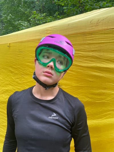 The media file shows a close-up, sideways shot of a young person wearing a purple helmet and bright green goggles. They are looking upwards and to the left, with their mouth slightly open. The person is wearing a black long-sleeved shirt with a white logo that reads "DEMIX" on the right side of the chest. The background is a crinkled yellow fabric, likely a tarp or tent. To the left of the yellow fabric, blurred green foliage is visible, suggesting an outdoor setting. The lighting appears to be natural, possibly indicating daytime. The overall impression is of someone preparing for or participating in an outdoor activity, perhaps camping or a sport that requires protective gear. The person's expression could be interpreted as one of anticipation or concentration.