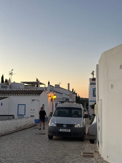 Here is a description of the image:
The image shows a street scene in Vila Real de Santo António, Portugal. A light gray Volkswagen van is parked on the right side of a narrow cobblestone street. The buildings on either side of the street are white with blue accents, typical of the region. The sky is a gradient of blue and orange, suggesting either dawn or dusk. A man in a dark-colored t-shirt and shorts is walking on the sidewalk next to the van. A street lamp is visible above one of the buildings, emitting a warm light.
