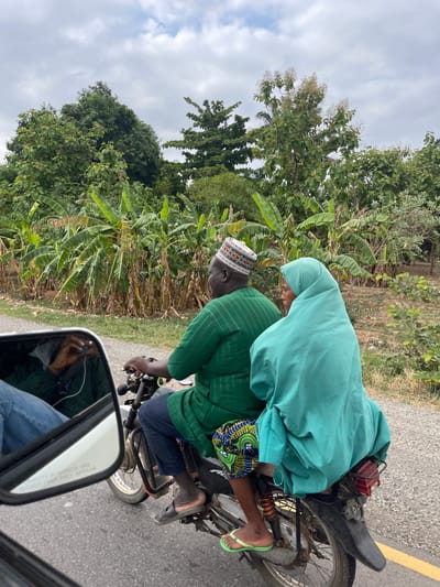 The image shows two people riding a motorcycle on a paved road in what appears to be a rural area in Mayo, Nigeria. The driver is wearing a green striped shirt, dark blue pants, a traditional hat, and is barefoot. The passenger is wearing a light blue head covering or hijab and brightly colored patterned shorts, and green flip flops. The motorcycle is black. Vegetation, including banana trees, grows along the side of the road. A car's side mirror is visible in the foreground, reflecting a person inside the car. The sky is overcast.