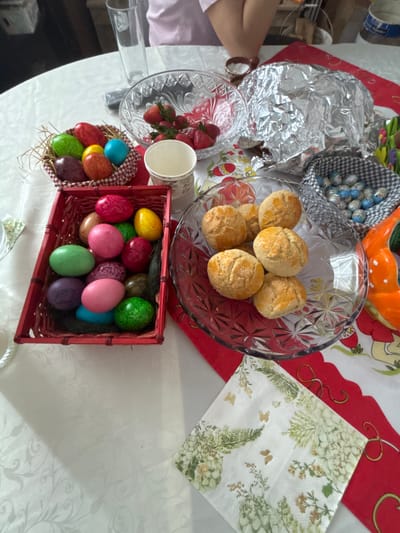 The image shows a table in Novo Panicharevo, Bulgaria, covered with a white patterned tablecloth and a red and white decorative runner. There are several bowls and containers with food items arranged on the table. A glass bowl contains a cluster of round, golden-brown baked goods. Next to it is a bowl filled with foil-covered objects and a small basket with blue-and-white patterned eggs. Another glass bowl is filled with fresh strawberries, with a stack of white paper cups next to it. A red basket contains a collection of colorful, dyed Easter eggs. A napkin with a floral and butterfly design is placed near the edge of the table. A person's arm and hand are visible at the edge of the table, wearing a bracelet.