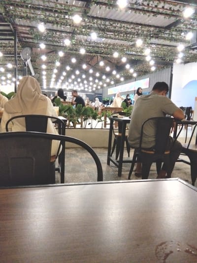 The image depicts an indoor dining area, possibly a restaurant or cafe, in Dalam, Indonesia. The ceiling is adorned with artificial greenery and numerous string lights, creating a festive atmosphere. In the foreground, a wooden table occupies the lower portion of the frame. In the mid-ground, a person wearing a beige hijab sits facing away from the camera. To the right, a man in a grey shirt is seated at a table, also facing away. Several other patrons are visible in the background, some wearing headscarves. Tables and chairs are arranged throughout the space. A large fan is mounted on a pole on the left side of the image. The overall ambiance suggests a casual dining establishment.
