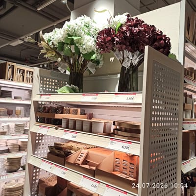 The image shows a retail store interior, likely a home goods or department store, in Modugno, Italy. The scene features shelves stocked with various products. In the foreground, there are displays of candles and decorative items. To the left, shelves are filled with stacks of plates and bowls. In the background, artificial flowers are displayed in vases. A price tag indicates "PREZZO BASSO SEMPRE" which translates to "ALWAYS LOW PRICE." The timestamp "24.07.2026 10:04" is superimposed on the lower right of the image. There are no people visible in the scene.